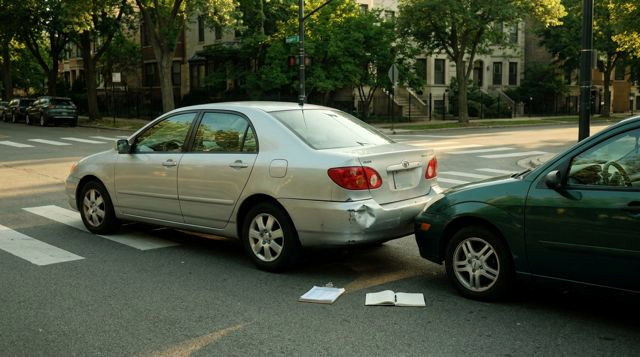 Silver sedan with a dented rear bumper stopped at a city crosswalk, a green car close behind it